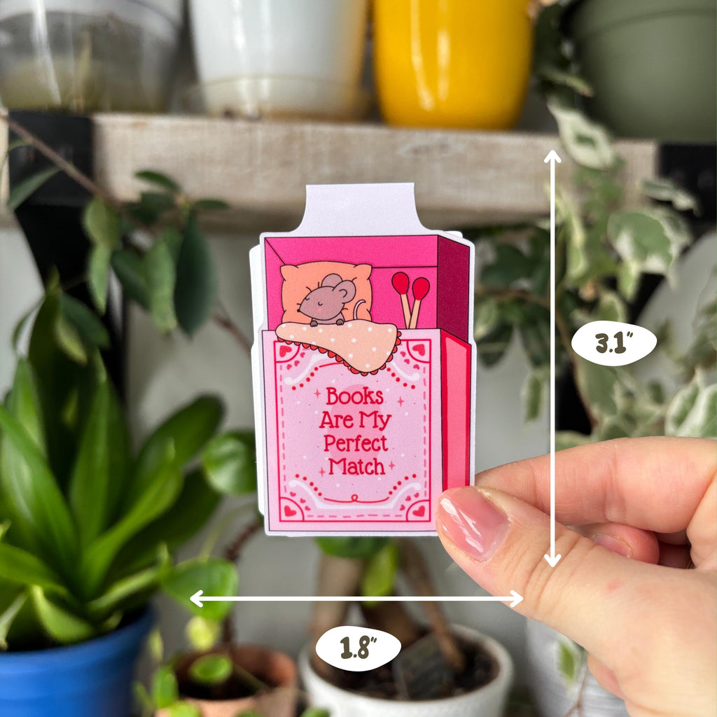 Small pink box with 'Books Are My Perfect Match' text held by a hand in front of potted plants.