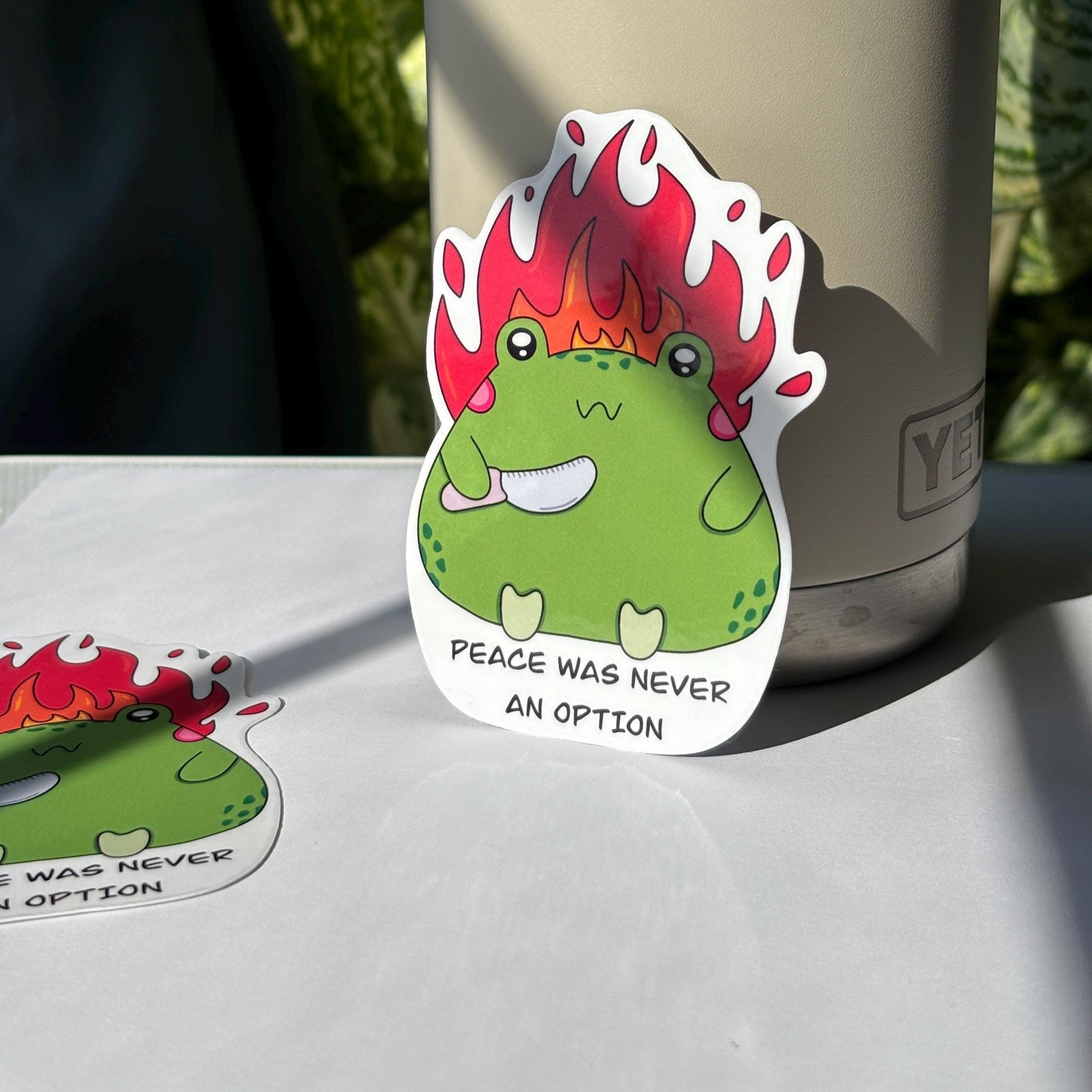 Sticker with a green creature on fire and text 'Peace was never an option' on a brown surface with a notebook.
