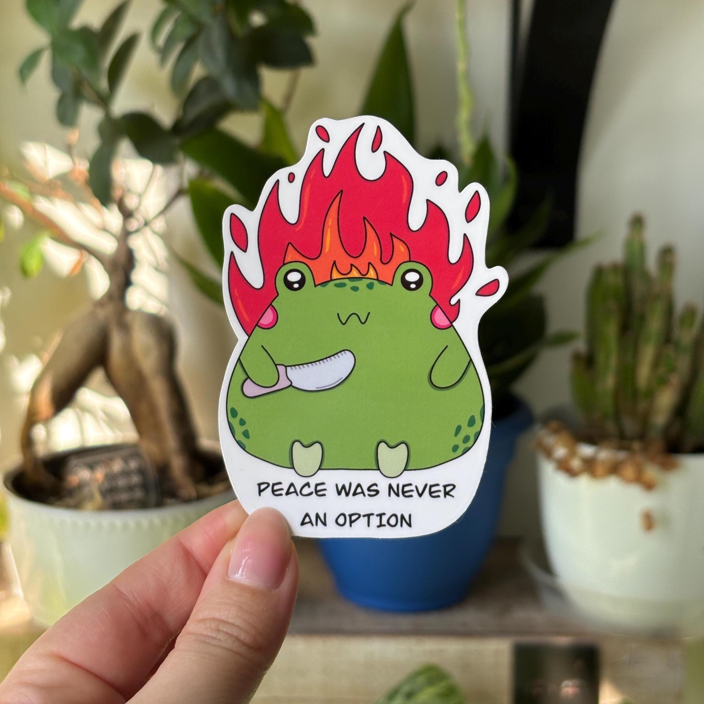 Hand holding a sticker with a green frog holding a knife with fire behind it and text 'Peace was never an option' underneath