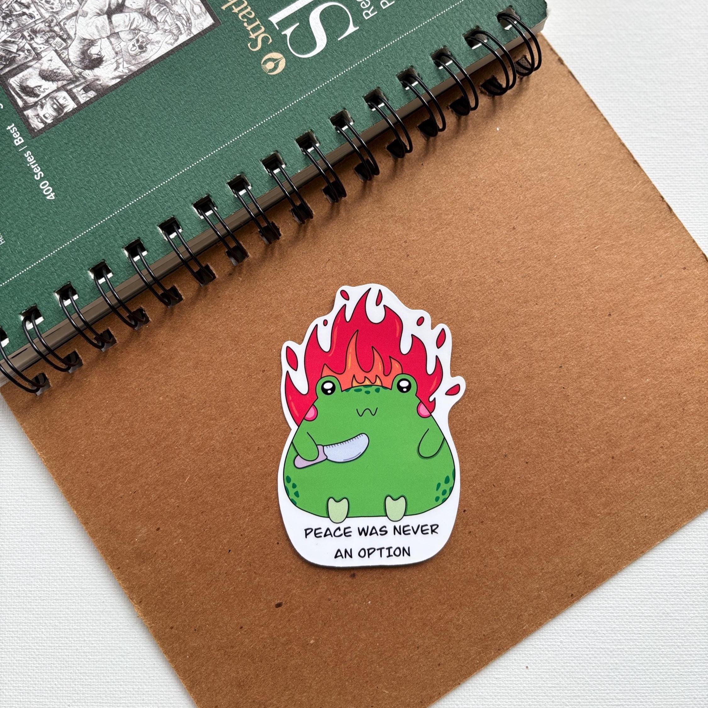 Sticker with a green creature on fire and text 'Peace was never an option' on a brown surface with a notebook.